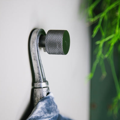 Schwarzer Messing-Möbelgriff von Brut Homeware als Wandhaken auf grauer Wand an dem eine Jacke mit Kleiderbügel hängt. Hintergrund ist Pflanzengrün.